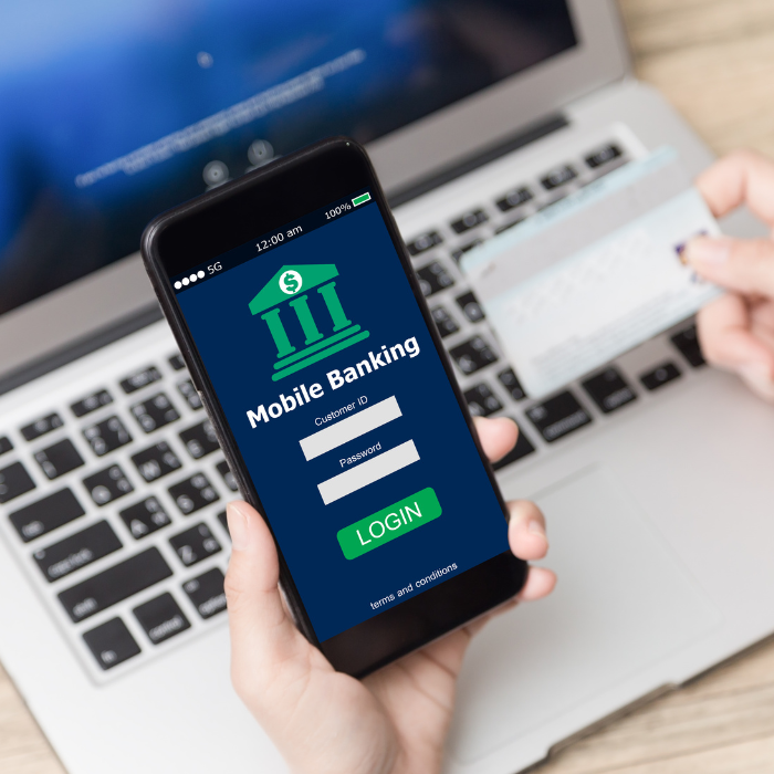 a hand holds a phone with mobile banking on the screen with a laptop underneath
